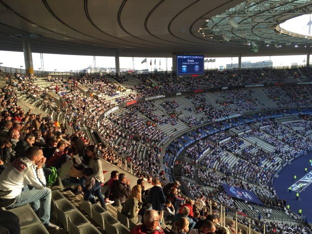 Große Menschenmenge in einem Stadion bei einem Fussballspiel sitzend, mit einer Bühne auf der rechten Seite, Fahnen, Masten, einem Bildschirm und der Allianz Arena in München, Deutschland im Hintergrund.