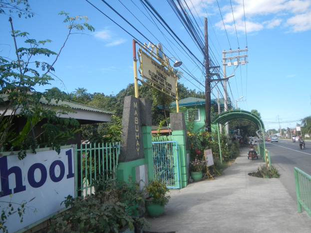 Einfahrt zu einer Schule mit einem "Schule"-Schild, Fahrzeuge auf der Straße, ein Tor, ein Tor, eine Tafel, Pflanzen, Bäume, Strommasten mit Drähten und ein bewölkter Himmel im Hintergrund.