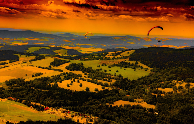 Zwei Paraglider über einem grünen Feld mit Bäumen, Häusern und Bergen im Hintergrund bei einer bewölkten Himmel.