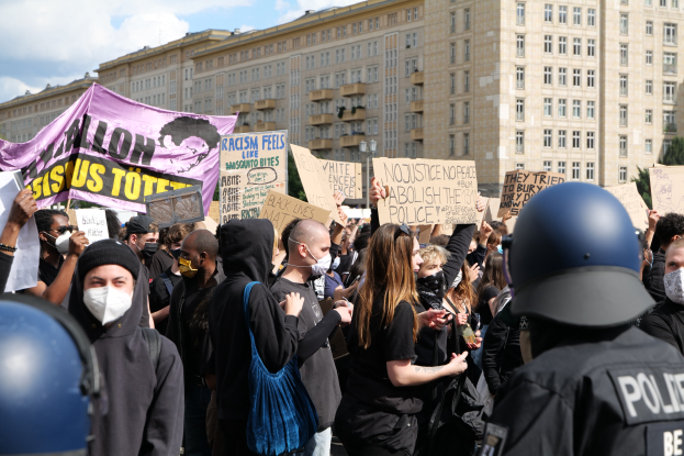 Gruppe von maskierten Menschen mit Schildern während einer Demonstration mit zwei Polizisten in Helmen auf der rechten Seite, Bäumen und bewölktem Himmel im Hintergrund.
