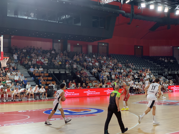Eine Gruppe von Menschen spielt Basketball in einer Turnhalle, mit einem Basketballkorb auf der linken Seite und Zuschauern auf Stühlen im Hintergrund, unter hellen Stadionlichtern.