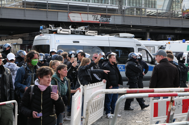 Eine Gruppe von Menschen steht vor einer Reihe von Polizeiwagen, einige tragen Helme und halten Telefone, mit Barrieren im Vordergrund und einer Brücke mit Geländern und Gebäuden im Hintergrund in Berlin, Deutschland.