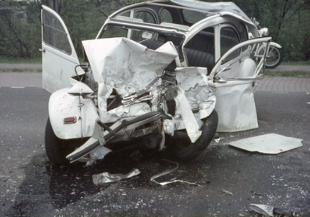Ein verunfalltes Auto mit schwerer Frontschaden am Straßenrand, mit Bäumen, einem Gebäude und einem anderen Fahrzeug im Hintergrund.