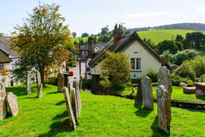 Ein kleines Dorf mit einem Friedhof im Vordergrund, umgeben von Gras, Steinen, Pflanzen, Bäumen, Häusern, Pfählen, Drähten und Fahrzeugen auf der Straße, mit Hügeln und einem bewölkten Himmel im Hintergrund und einem prominenten Baum in der Mitte.