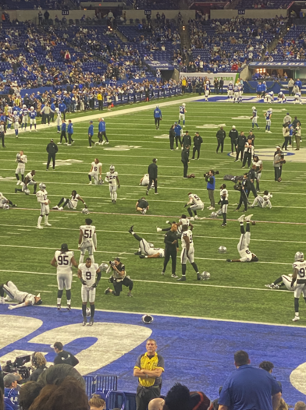 Ein Fußballspiel wird in einem Stadion mit Menschen auf dem Rasen und einigen auf Stühlen im Hintergrund sowie einer Person mit einer Kamera gespielt, was auf eine Live-Übertragung hinweist.