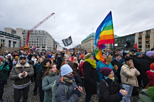 Eine große Gruppe von Menschen bei einer LGBTQ+-Rechtsmarsch in Berlin, die Fahnen und Banner schwenken, mit Gebäuden und einem Kran im Hintergrund unter einem bewölkten Himmel.