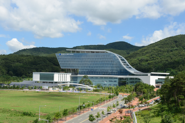 Großes modernes Gebäude mit vielen Fenstern, als Hauptsitz der Korea National University of Science and Technology bezeichnet, in einer parkähnlichen Landschaft mit Bäumen, Gras und einer Straße mit Fahrzeugen umgeben, von Hügeln umgeben unter einem bewölkten Himmel.