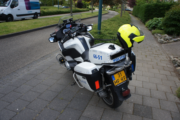 Ein Polizei-Motorrad am Straßenrand mit einem Helm darauf, umgeben von Bäumen, Pfählen, Gebäuden, Pflanzen, Gras, Steinen und anderen Fahrzeugen im Hintergrund.