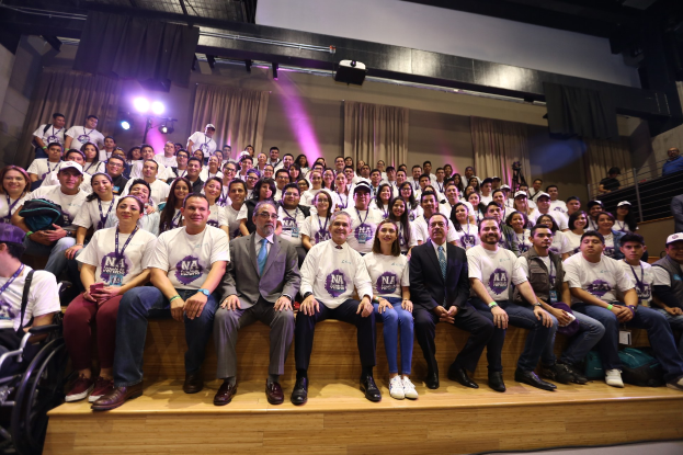 Eine Gruppe von Menschen sitzt auf einer Bühne, wobei sich links ein Mensch im Rollstuhl befindet, bei einem Event mit Vorhängen, Lichtern und einer Wand.
