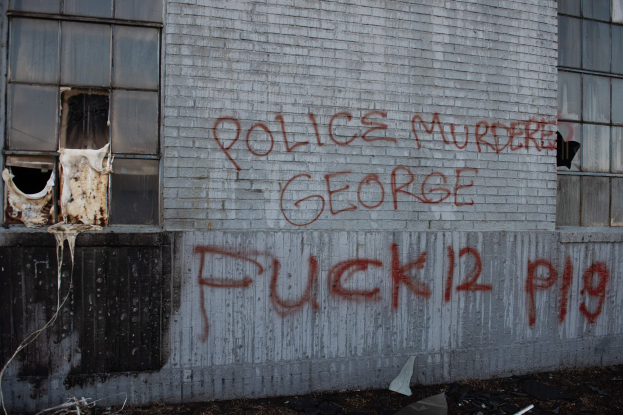 Graffiti an einer Gebäudewand mit der Aufschrift 'Polizei Mord George Fuck 12 P19' und Fenstern sowie verstreuten Gegenständen davor.