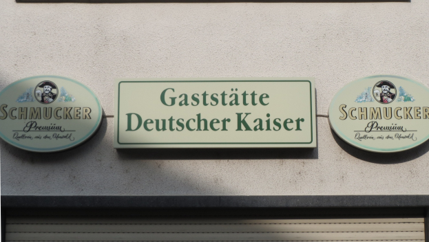 Drei hölzerne Bretter mit der Aufschrift "Gaststätte Deutscher Kaiser Schmucker" sind an einer Gebäudewand über einer geschlossenen Rolllade angebracht.