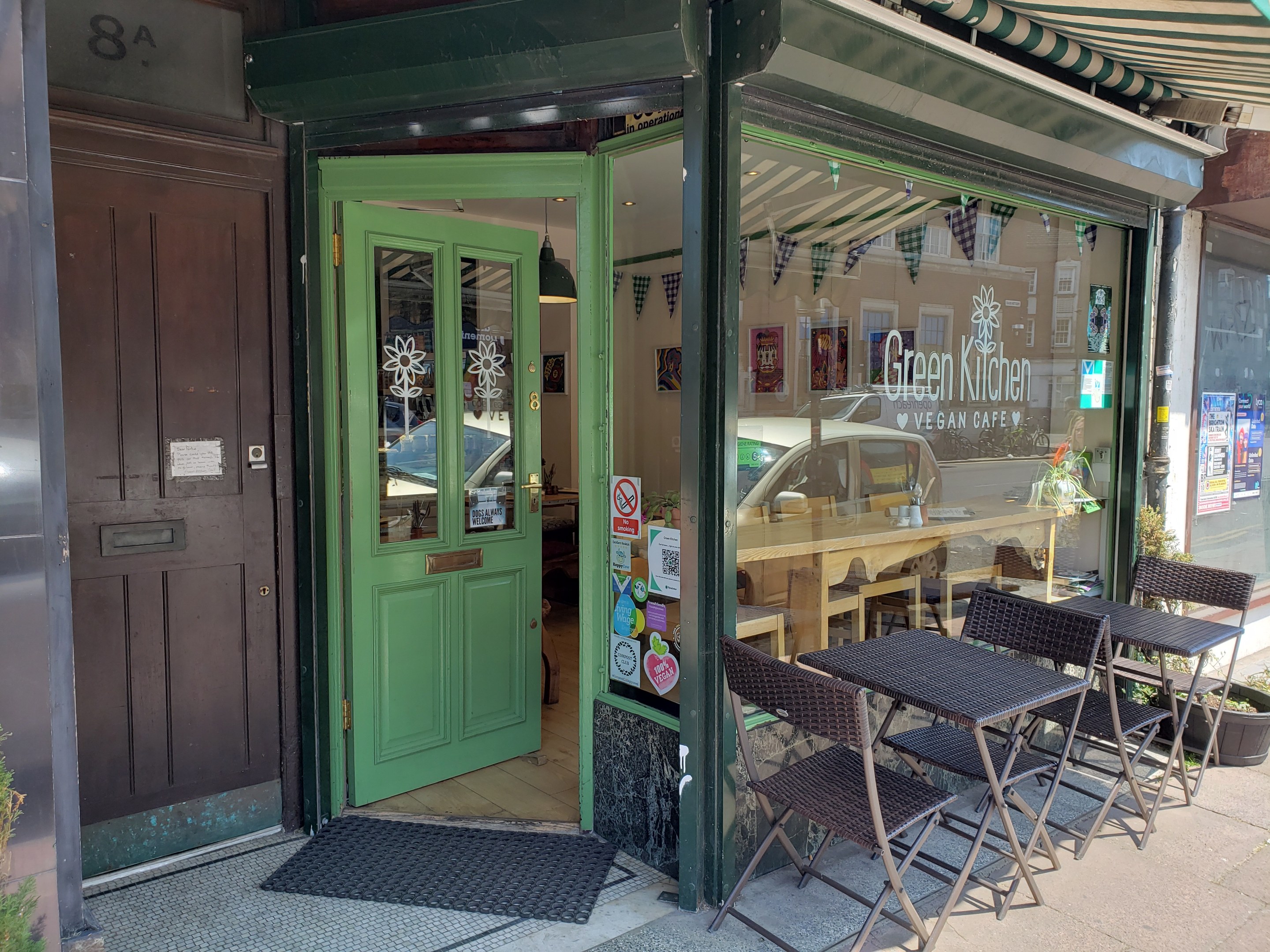 Ein Restaurant namens Green Kitchen Vegan Cafe mit Außenbereich, umgeben von Pflanzen, mit einer Glaswand voller Plakate, einer Fußmatte und Innenmöbeln und Beleuchtung.