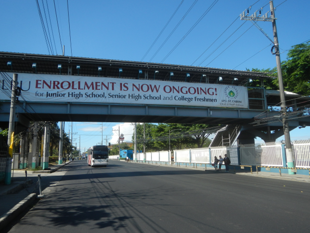 Ein Bus fährt unter einer Brücke mit einem Schild, auf dem "Jetzt Anmeldung" steht, vorbei, mit Menschen, die auf dem Gehweg darunter laufen, Strommasten mit Drähten entlang der Straße und Bäumen mit einem klaren blauen Himmel im Hintergrund.