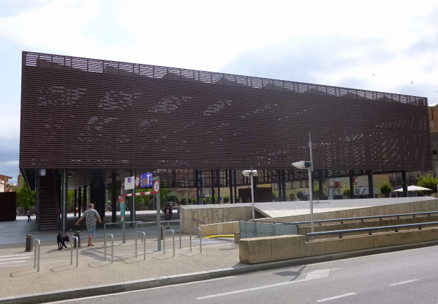 Großes modernes Gebäude mit zahlreichen Fenstern, das als neuer Eingang zur Stadtmitte dient, flankiert von Straßeninfrastruktur und Fußgängern.