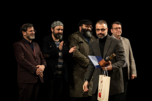 Eine Gruppe Männe steht auf einer Bühne, einer hält eine Tasche und eine Trophäe, mit einem dunklen Hintergrund, der die Eröffnung der Berliner Filmfestspiele suggeriert.