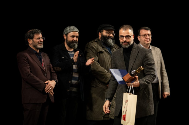 Eine Gruppe Männe steht auf einer Bühne, einer hält eine Tasche und eine Trophäe, mit einem dunklen Hintergrund, der die Eröffnung der Berliner Filmfestspiele suggeriert.