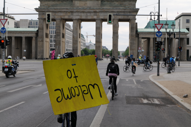 Eine Gruppe von Radfahrern fährt an der Brandenburgertor in Berlin vorbei, einer hält ein gelbes Schild, mit Laternenmasten, Verkehrszeichen, Gebäuden, Bäumen und einem klaren Himmel im Hintergrund.