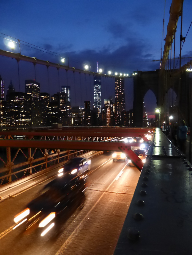 Ein Auto fährt nachts über die Brooklyn Bridge, mit beleuchteten Gebäuden und Sternen im Hintergrund.