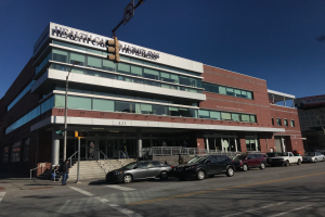 Ein großes Gebäude mit parkenden Autos davor, umgeben von Straßenlaternen, Lampen, Verkehrszeichen, Menschen auf dem Gehweg, Geländern, Treppen, Bäumen und einem klaren blauen Himmel, identifiziert als das New York City Health Care Homeless Center.