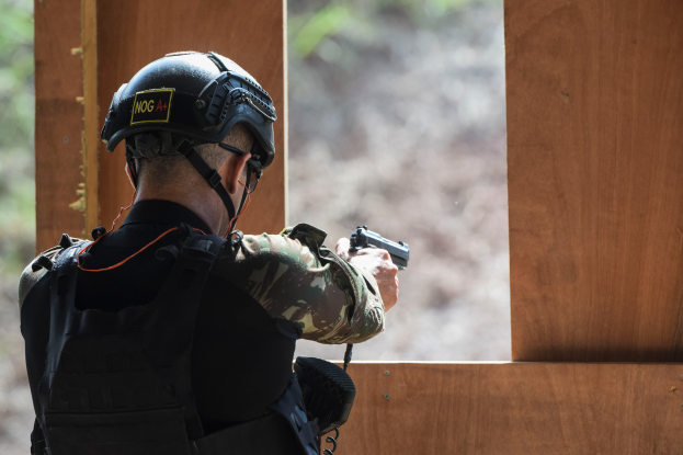 Ein Mann in Tarnkleidung, schwarzer Jacke und Helm zielt mit einer Pistole auf ein Ziel, mit einem unscharfen Hintergrund.