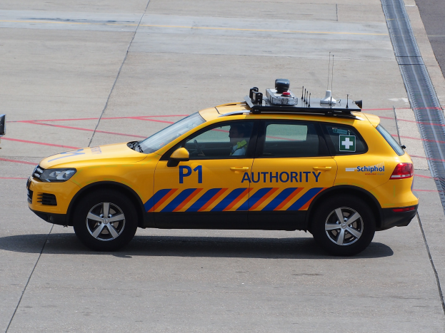 Ein gelbes Fahrzeug, das als Taxi gekennzeichnet ist, das auf dem Rollfeld eines Flughafens geparkt ist, mit einer Person darin und einem anderen Fahrzeug, das teilweise links zu sehen ist.