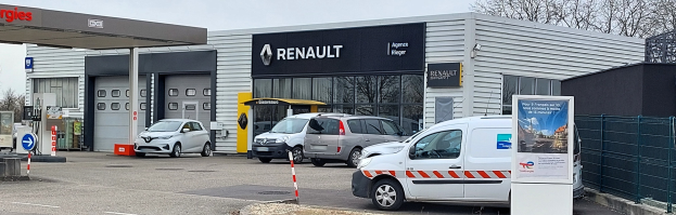 Renault-Autohaus mit parkenden Autos vorne, umgeben von einem Metallzaun, einer Texttafel, Pfosten, einem Schild, Bäumen und einem bewölkten Himmel.