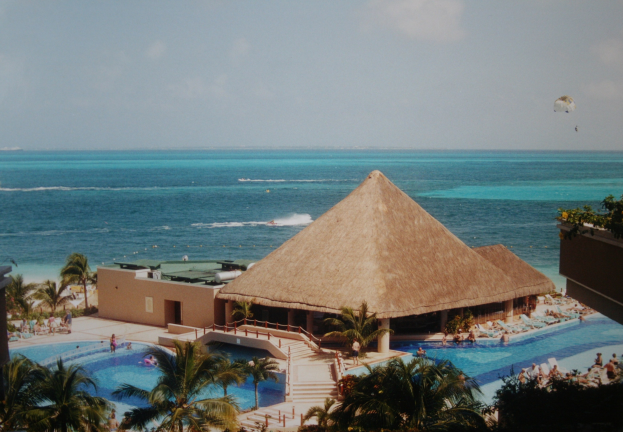 Resort mit einem gro├čen Schwimmbad umgeben von Palmen, Hütten und Menschen, mit einem Ozean, bew├Âlktem Himmel und einem Fallschirm im Hintergrund.
