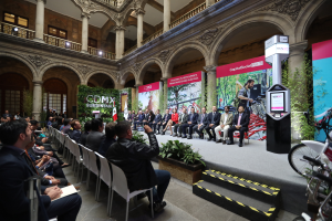 Eine Gruppe von Menschen sitzt auf Stühlen und schaut auf einen Mann auf einer Bühne, der ein Mikrofon hält, rechts stehen Fahrräder und im Hintergrund sind Plakate, Pflanzen, Säulen, Geländer, Lampen, Fenster und eine Wand zu sehen.