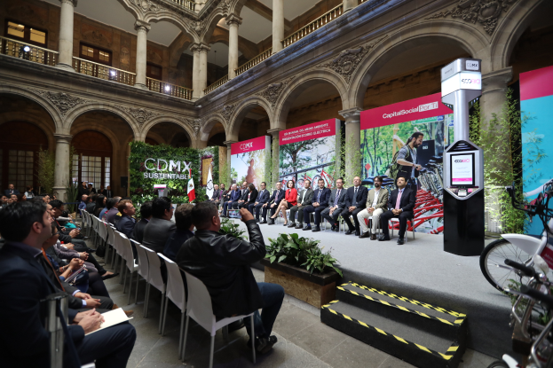 Eine Gruppe von Menschen sitzt auf Stühlen und schaut auf einen Mann auf einer Bühne, der ein Mikrofon hält, rechts stehen Fahrräder und im Hintergrund sind Plakate, Pflanzen, Säulen, Geländer, Lampen, Fenster und eine Wand zu sehen.