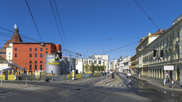 Stadtstra√enbild mit Gebäuden, Fahrzeugen, Passanten und Infrastruktur unter einem Himmelhintergrund.