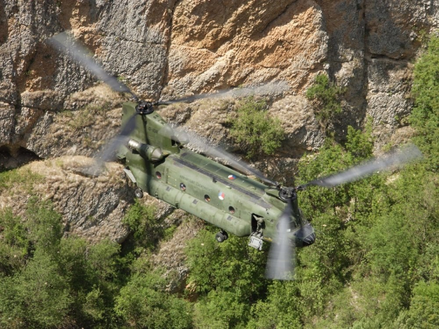 Ein Militärhubschrauber fliegt über eine felsige Klippe mit Bäumen im Vordergrund.