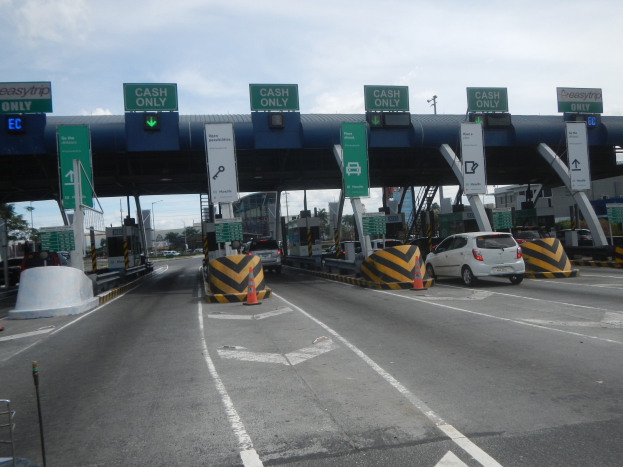 Mautstelle mit darunterfahrenden Autos, umgeben von Verkehrskegeln, Absperrpfosten, Schildtafeln, einem Torbogen, Laternenmasten, B├Ąumen, Geb├Ąuden und einem bew├Âlktem Himmel.