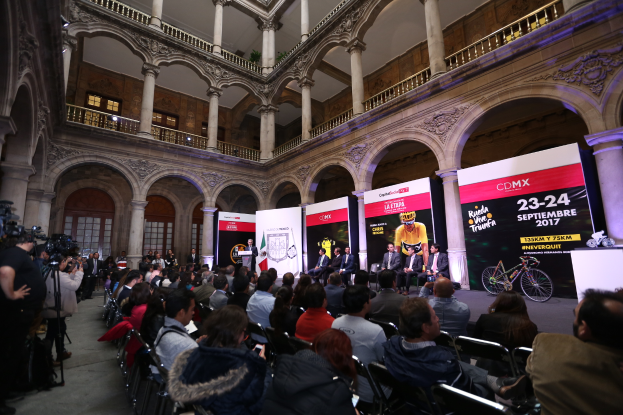 Eine Gruppe von Menschen, die an einem Tisch mit einem Podium und einem Mikrofon vor einer Menge, Kameras und Transparenten sitzt, was auf eine Pressekonferenz für das CDMX 2017-Event hindeutet.