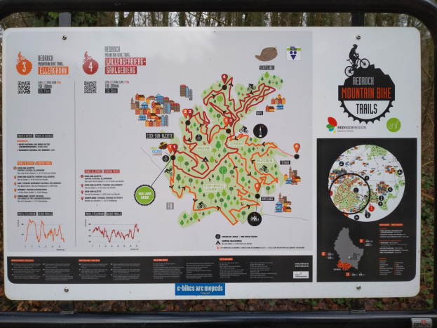 Ein Wegweiser mit einer Karte einer Mountainbike-Strecke in einem Waldgebiet, an einem Metallstab befestigt, unter einem klaren blauen Himmel.