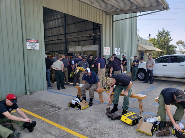 Eine Gruppe von Menschen in Militäruniformen nimmt an einer Outdoor-Trainingseinheit teil, bei der verschiedene Ausrüstungsgegenstände, Fahrzeuge und Gebäude im Hintergrund zu sehen sind.