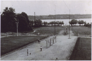 Ein Schwarz-Weiß-Foto von Menschen, die Fußball spielen, umgeben von Bäumen, Pfählen und einem Zaun, mit einem Wasserlauf und einem klaren Himmel im Hintergrund und Text unten.