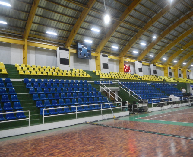 Großer Hallensportplatz mit Holzboden, blauen und gelben Sitzplätzen, Geländern, Treppen, wandmontierten Bildschirmen, Deckenleuchten und einigen Personen.