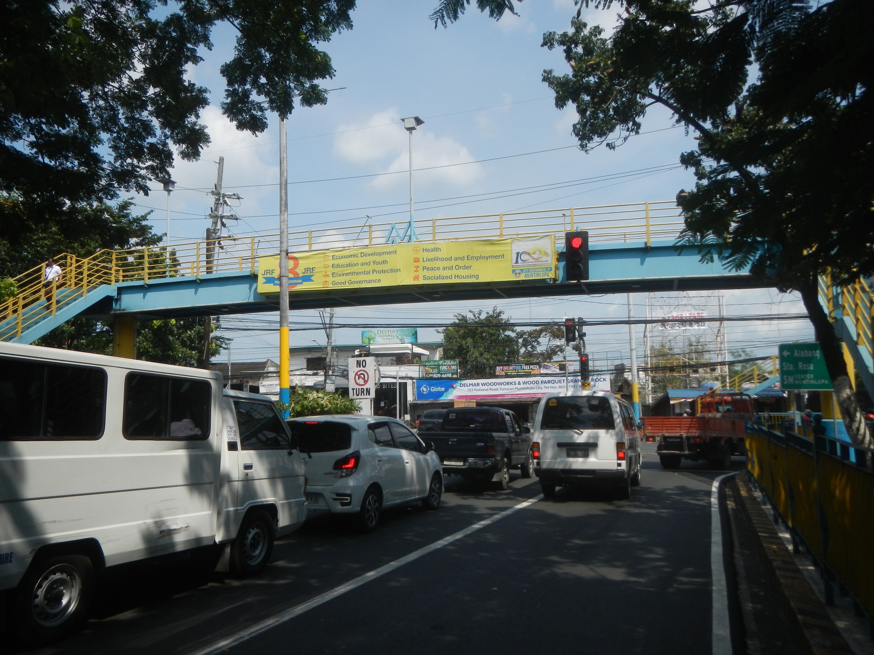 Vielbefahrene Straße mit Fahrzeugen, eine Brücke mit Geländern und Treppen, Laternen, Verkehrsampeln, Texttafeln, Bäume, Gebäude und ein bewölkter Himmel.