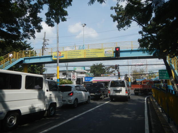 Vielbefahrene Straße mit Fahrzeugen, eine Brücke mit Geländern und Treppen, Laternen, Verkehrsampeln, Texttafeln, Bäume, Gebäude und ein bewölkter Himmel.