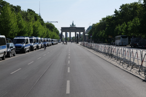Lange Reihe von Polizeiwagen auf der Straße vor dem Brandenburger Tor in Berlin geparkt, mit Menschen auf Fahrrädern und in der Nähe stehenden, Barrieren, Bäumen, einem Bogen mit Statuen im Hintergrund und sichtbarem Himmel.