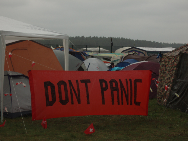 Gruppe von Zelten mit einer roten Fahne, auf der 'Don't Panic' steht, im Vordergrund, umgeben von Gras und Fahnen, mit Bäumen und einem klaren blauen Himmel im Hintergrund.