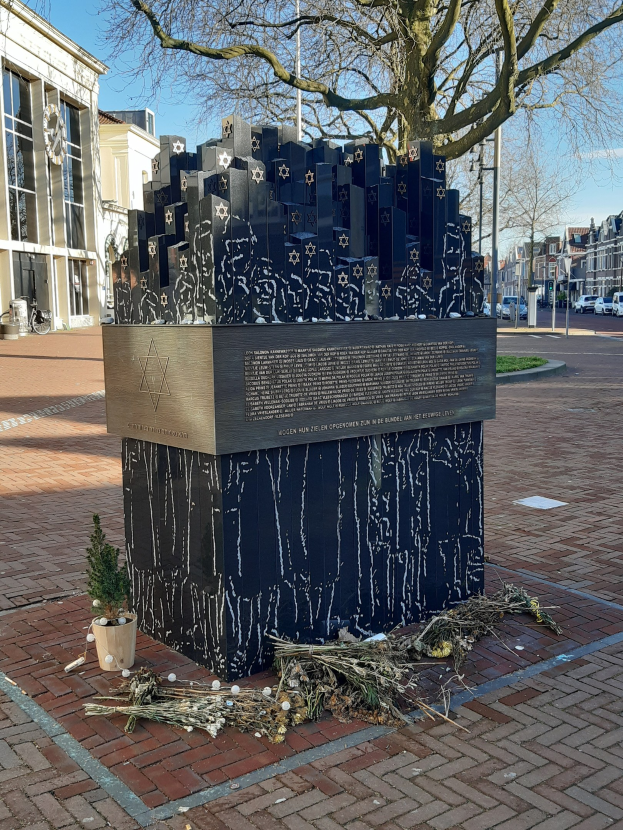 Denkmal in einem Stadtplatz für Holocaust-Opfer mit einem Baum davor, einem Topfgewächser auf dem Boden, verstreuten trockenen Blättern und Gebäuden, Fahrzeugen, Masten und Bäumen im Hintergrund.