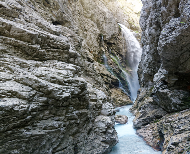 Ein kleiner Wasserfall stürzt sich über zerklüftete Felsen in einem steinigen Tal hinab, umgeben von saftig grünen Hügeln unter strahlendem Sonnenschein.
