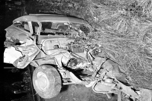 Ein Schwarz-Weiß-Foto eines schwerbeschädigten Autos am Straßenrand mit einer zerbeulten Front, zersplitterten Fenstern, einem Baum im Hintergrund und verstreuten Gegenständen um die Unfallstelle.