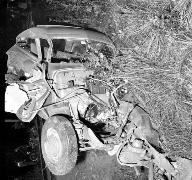 Ein Schwarz-Weiß-Foto eines schwerbeschädigten Autos am Straßenrand mit einer zerbeulten Front, zersplitterten Fenstern, einem Baum im Hintergrund und verstreuten Gegenständen um die Unfallstelle.