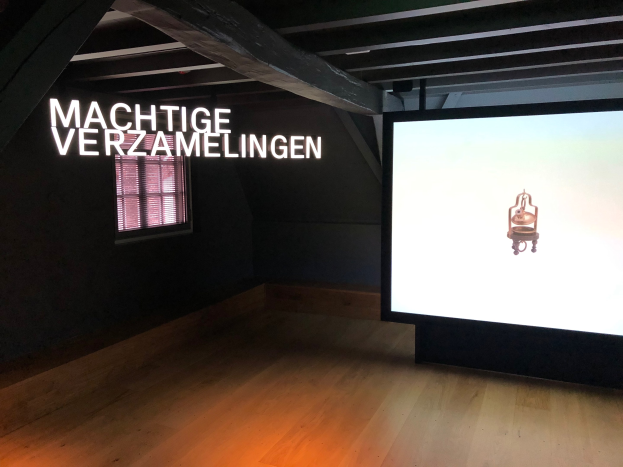 Installationsansicht der Ausstellung 'Machtige Verzamelingen' im Museum of Modern Art in Berlin mit einem großen Bildschirm an der Wand, einem Fenster links und sichtbarer Decke.