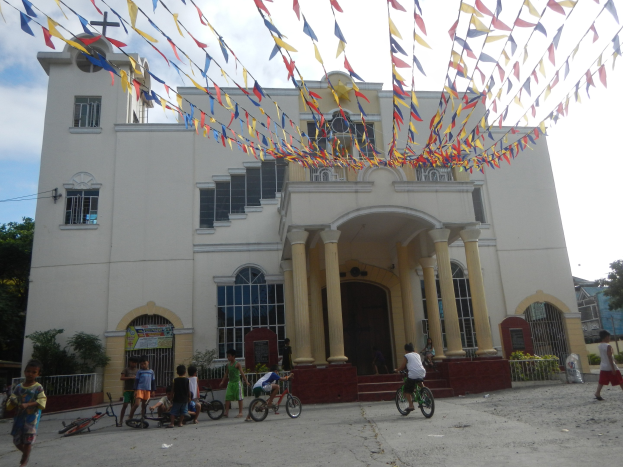 Eine Gruppe von Menschen auf Fahrrädern vor einer weißen Kirche mit architektonischen Details wie Fenstern, Säulen, Bögen, Türen und einem Kreuzsymbol, umgeben von Pflanzen, einem Zaun, einer Fahne, Bäumen und einem bewölkten Himmel, mit bunten Fahnen an einem Seil oben.