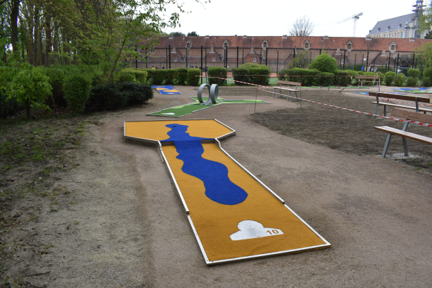 Minigolfanlage in Parkumgebung mit Bänken, Grünfläche, einem Zaun, Gebäuden im Hintergrund und einem klaren blauen Himmel.