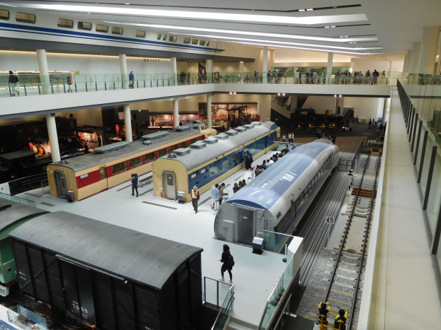 Eine großzügige Museumsausstellung zeigt verschiedene Züge in unterschiedlichen Größen und Farben, bei der Besucher von verschiedenen Ebenen und dem Erdgeschoss aus zuschauen.