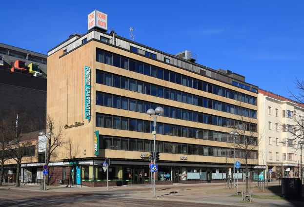 Großes Gebäude mit vielen Fenstern, als das Fore Inn Hotel Berlin identifiziert, umgeben von Straßeninfrastruktur einschließlich Pfosten, Lampen, Signalen, Schildern, Bäumen und einem bewölkten Himmel.